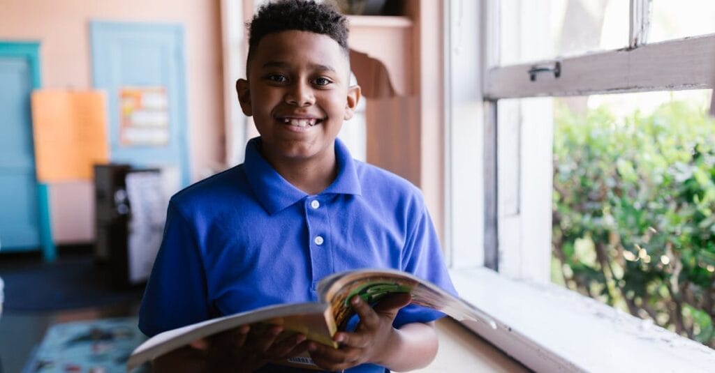 Young boy in blue polo reading by a classroom window, smiling happily.