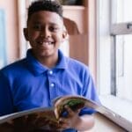 Young boy in blue polo reading by a classroom window, smiling happily.