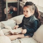 A cute little girl reading a book in a warm and cozy bedroom environment.