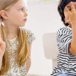 Two children sitting indoors engaging in a playful learning activity, showing numbers with fingers.