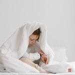 A young child sitting playfully under a white duvet on a bed in a bright room.