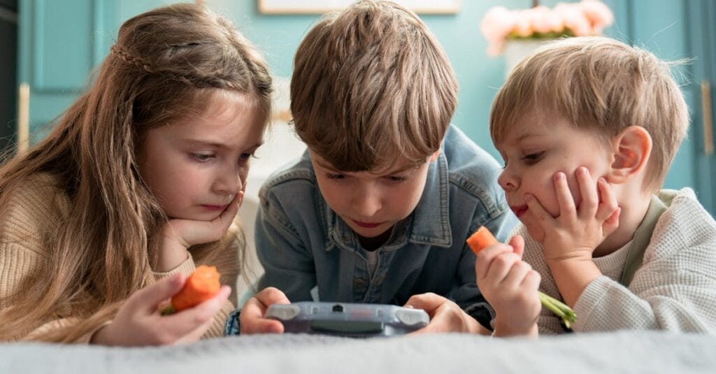 Three kids enjoying video games and snacking on carrots indoors, showcasing childhood togetherness.