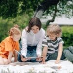 Three kids reading a book on a picnic blanket in a park under a tree.