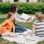 A woman teaching two children outdoors on a blanket, fostering education in nature.