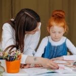 Teacher helps young girl with learning activity in a classroom setting, fostering education.