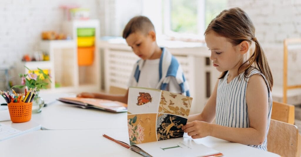 Two children reading books at a bright indoor classroom table filled with colorful supplies.