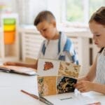 Two children reading books at a bright indoor classroom table filled with colorful supplies.