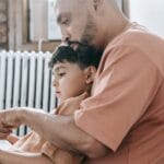 A father teaches his son to read, creating a warm bonding moment in a cozy home setting.