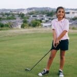 A smiling girl holding a golf club on an outdoor golf course, enjoying a sunny day.