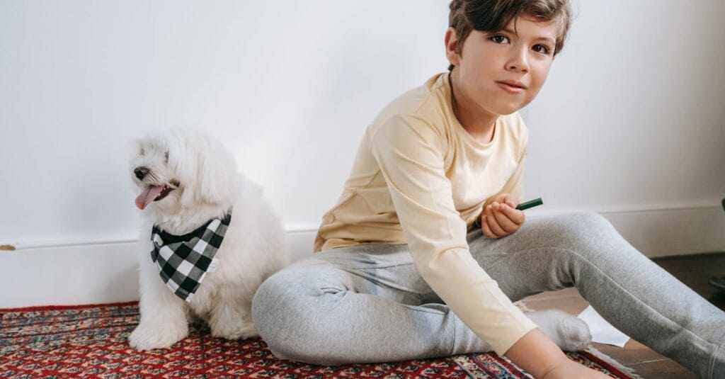 A young boy sitting indoors on a carpet playing with pencils beside a Maltese dog.