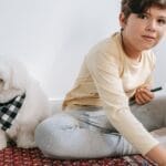 A young boy sitting indoors on a carpet playing with pencils beside a Maltese dog.