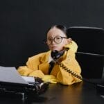 A child in a yellow suit sits at a desk with a phone, typewriter, and globe, portraying a business scene.