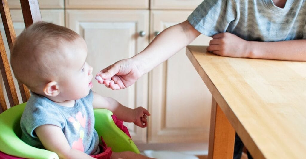 A cute baby in a high chair being fed by an older child in a bright kitchen setting.