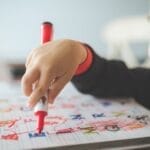 A young child using a marker to write letters in an educational workbook.