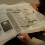 Person reviewing job listings in a newspaper, holding a pink marker for highlighting job ads.