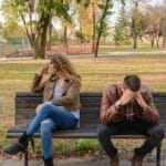 An upset couple seated on a park bench, expressing frustration during an autumn day.