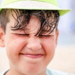 Close-up of a smiling boy with eyes closed and wearing a hat on a sunny beach day.