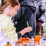A child enjoys painting pumpkins at an outdoor crafting event, surrounded by art supplies.