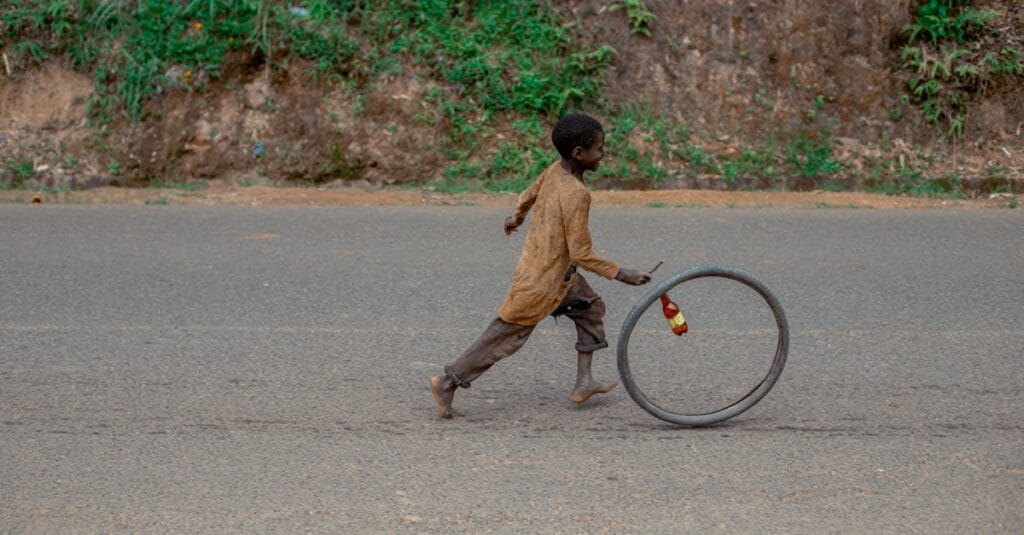 A child joyfully runs alongside a rolling bicycle wheel on a rural road, capturing a moment of simple play.
