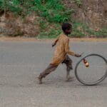 A child joyfully runs alongside a rolling bicycle wheel on a rural road, capturing a moment of simple play.