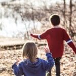Two children running through a forest path by a lakeside on a sunny day.
