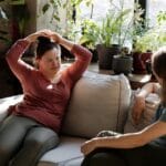 Two women enjoying a relaxed conversation on a sofa in a bright room filled with plants.