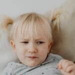 A young blonde child with a displeased facial expression on a light background.