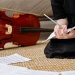 A young girl in a black dress sits on a woven carpet, writing music notes beside her cello in a cozy living room.