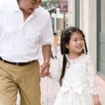 Elderly man and young girl holding hands and smiling while walking outdoors.